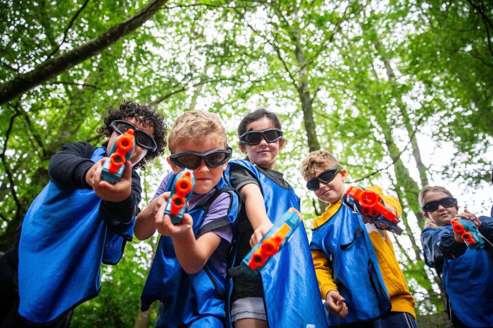 Five kids in blue vests aiming toy guns, wearing goggles, in a forest setting.