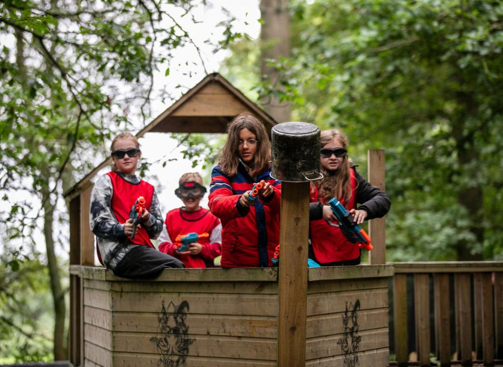 Four kids in red vests with toy guns play in a wooden fort in the woods.