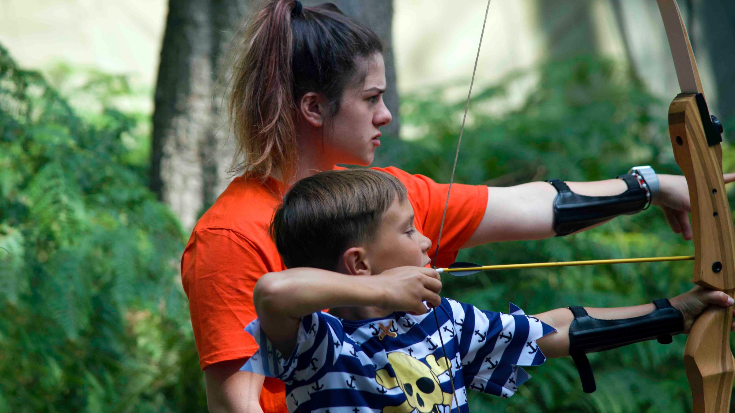 Woman teaching a young boy archery in an outdoor setting.