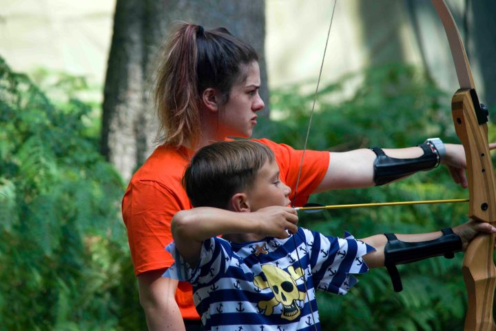 Woman teaching a young boy archery in an outdoor setting.