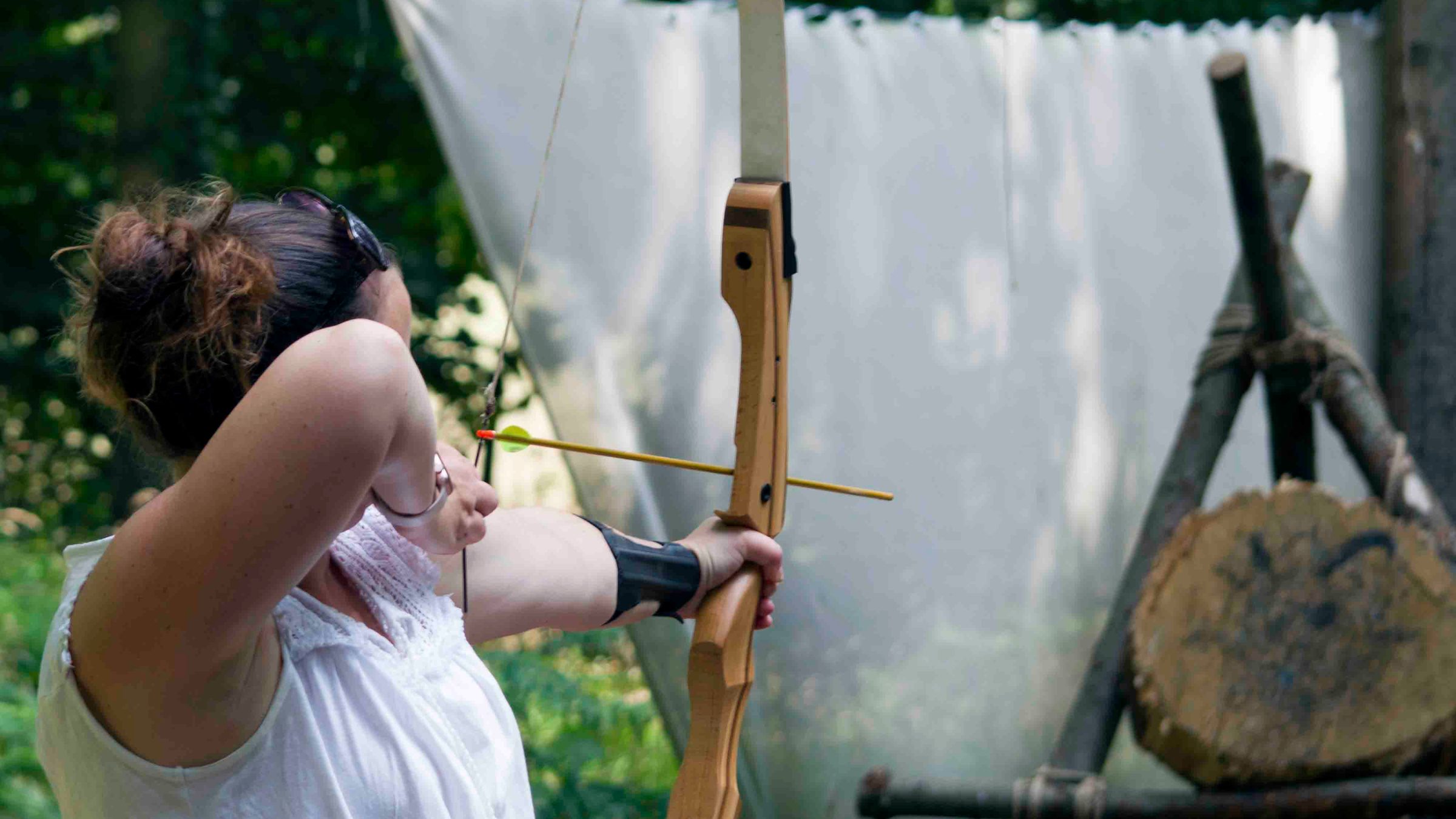 Person practicing archery outdoors, aiming a bow at tree stump targets.
