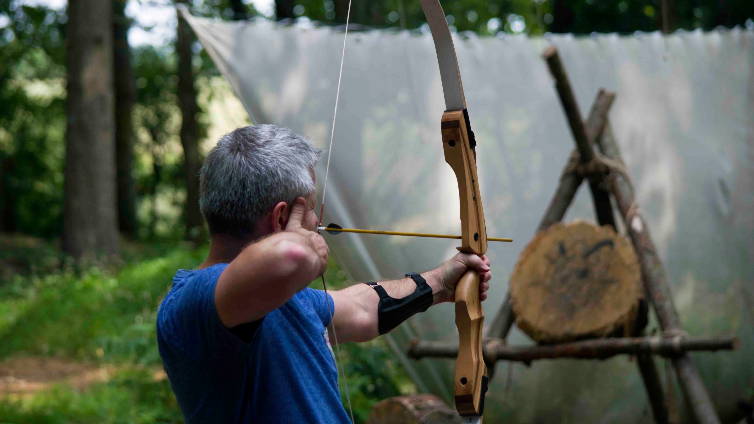 Person in blue shirt aiming a bow and arrow at a target in a forest setting.