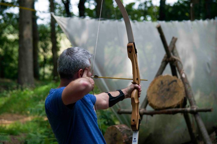 Person in blue shirt aiming a bow and arrow at a target in a forest setting.