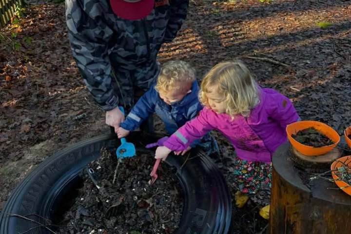 Two children digging in soil inside a tire with an adult in a forest setting.