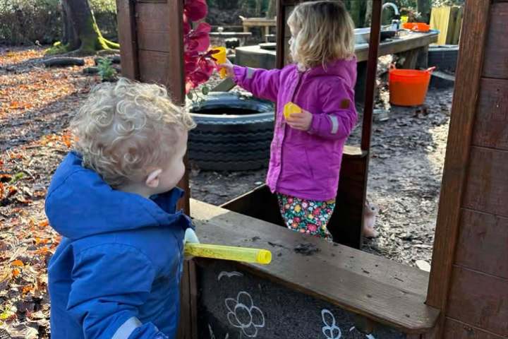 Two children playing at an outdoor playhouse labeled 'HOME' with leaves and chalk drawings.