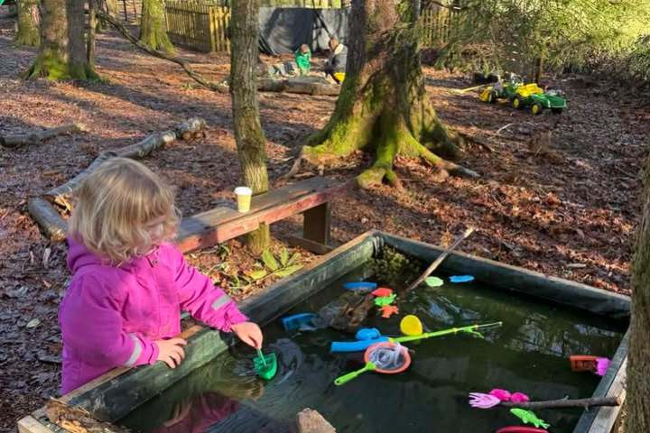 Child in pink jacket playing with toys in outdoor water table under trees.