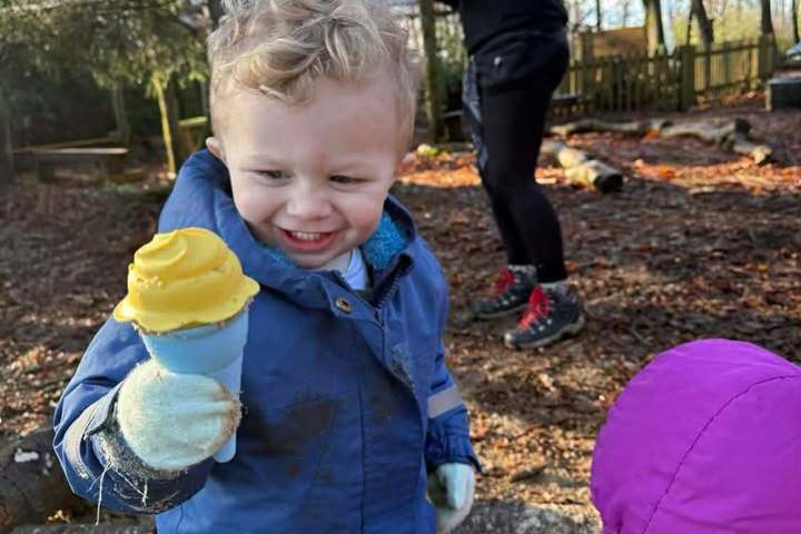 Child in blue coat holds toy ice cream cone in sandy playground with adults and another child nearby.