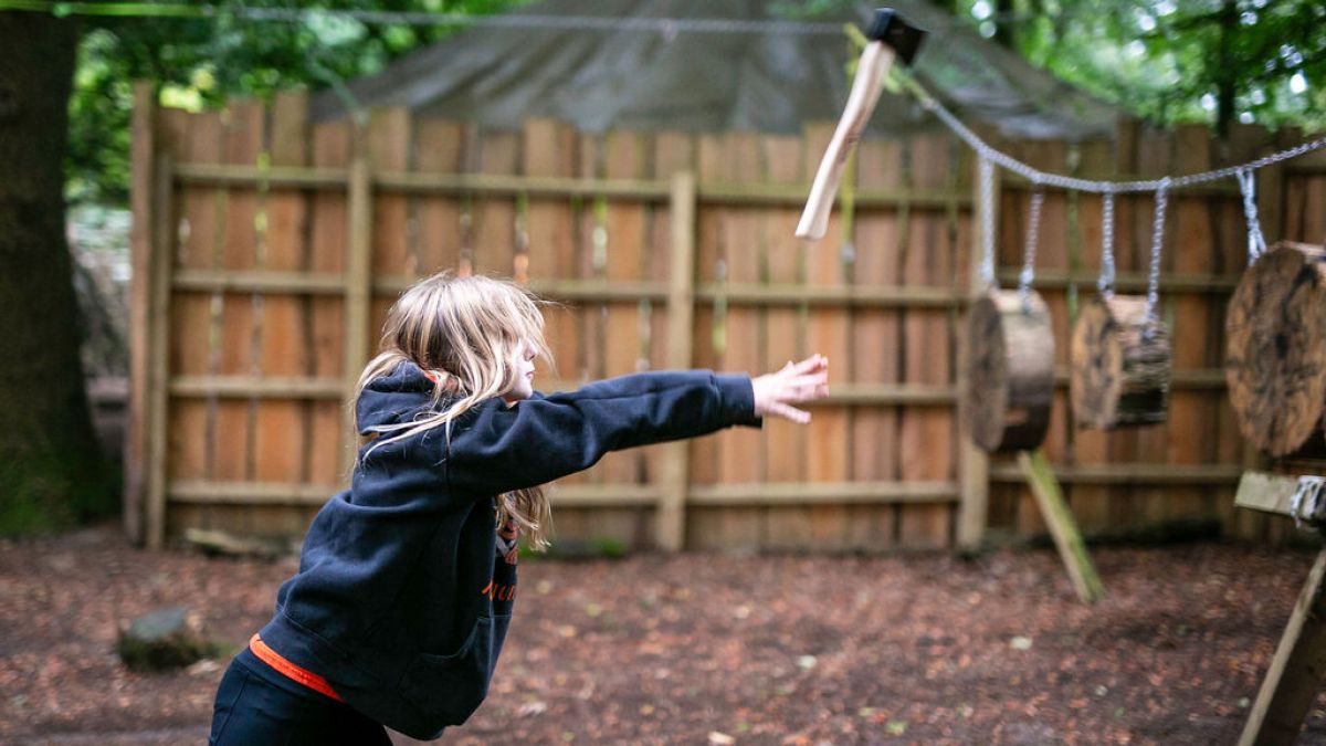 Child in hoodie throwing an axe at a wood target in a wooded area.