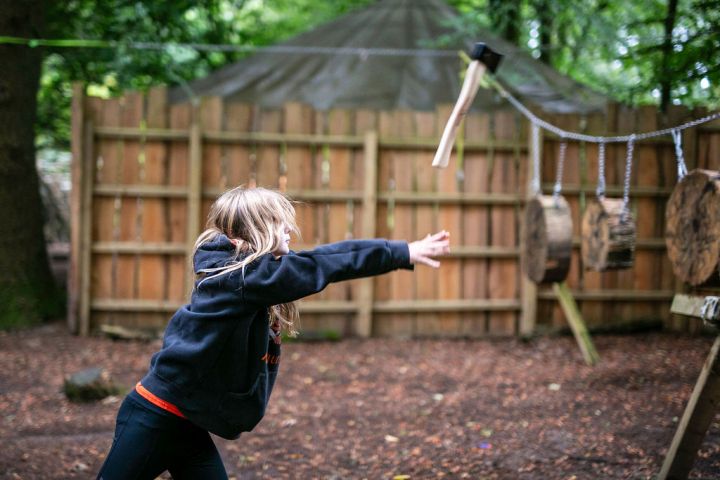 Child in hoodie throwing an axe at a wood target in a wooded area.