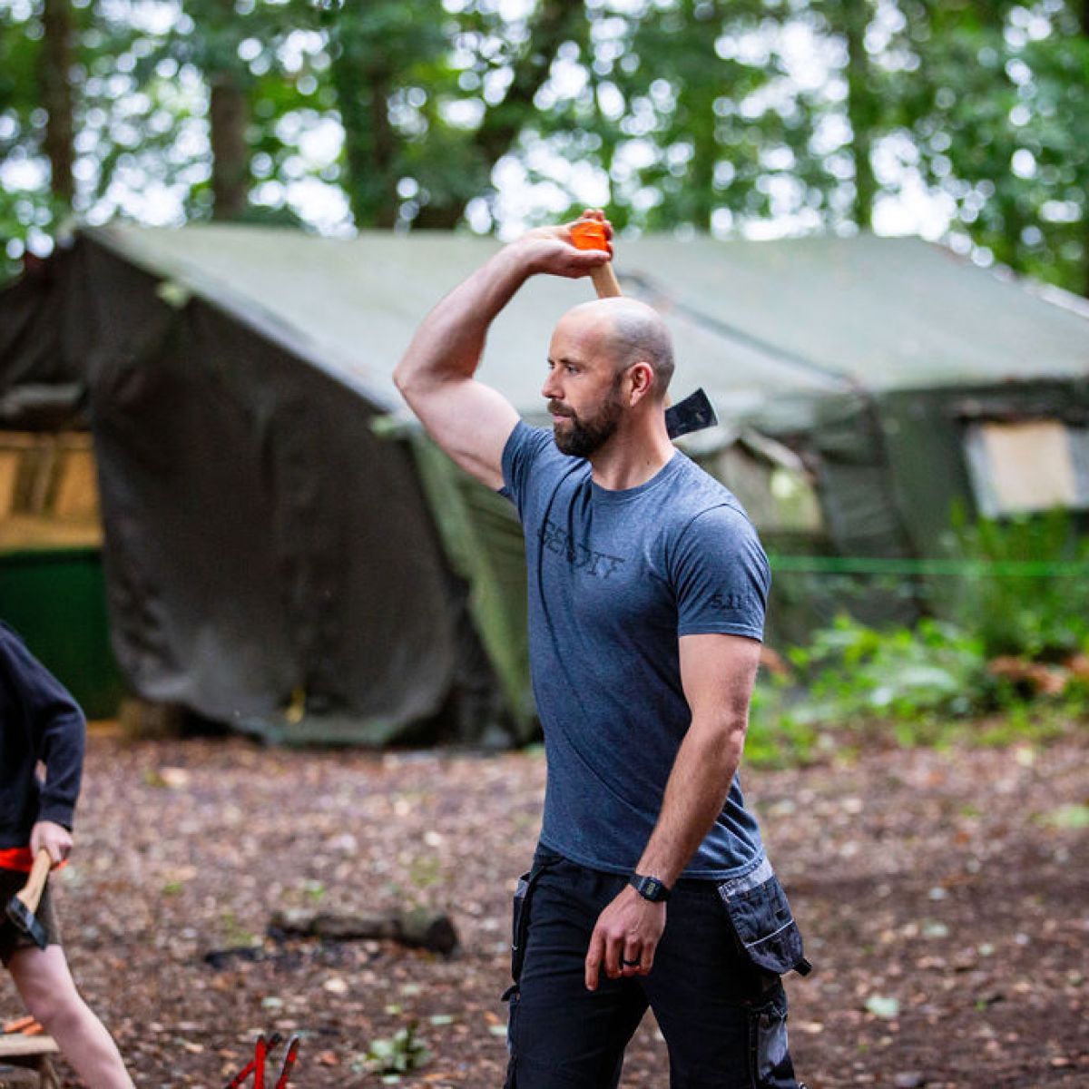 Man throwing an axe at outdoor camp, boy holding axe nearby, tent in background.