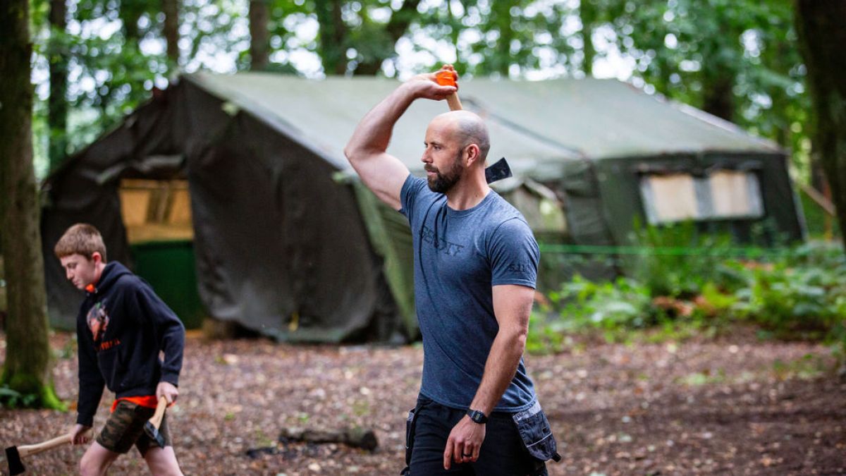 Man throwing an axe at outdoor camp, boy holding axe nearby, tent in background.