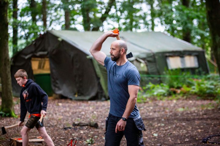 Man throwing an axe at outdoor camp, boy holding axe nearby, tent in background.