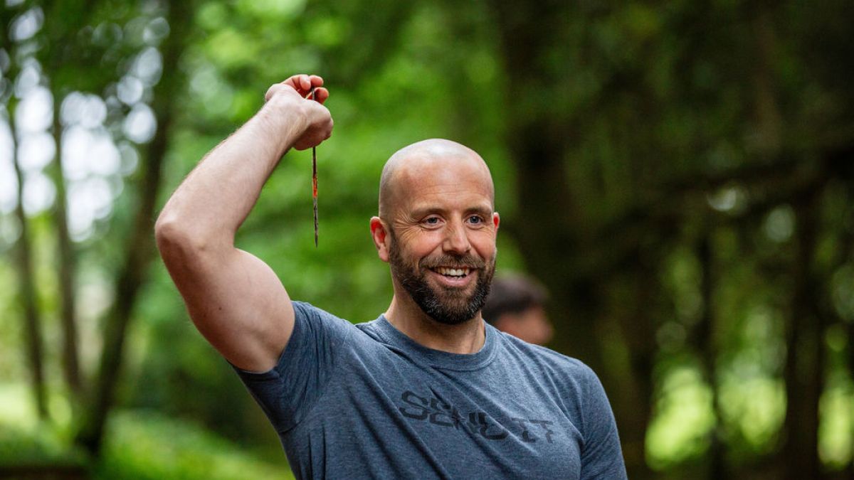 Man holding a knife, smiling in a forest setting.