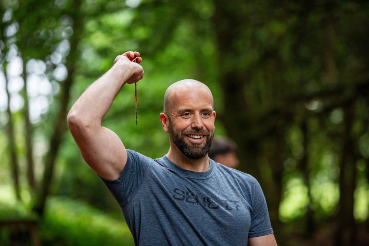 Man holding a knife, smiling in a forest setting.