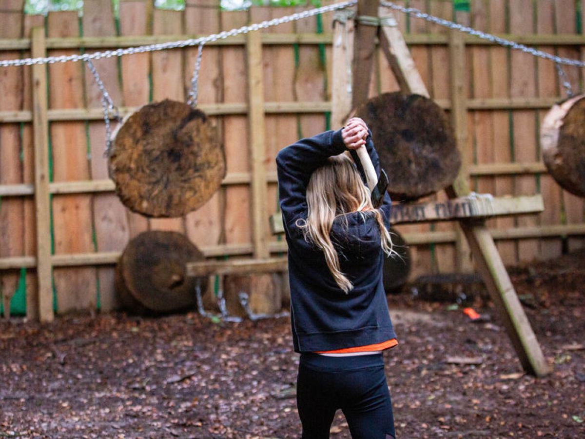 Person with long hair prepares to throw an axe at wooden targets in a fenced outdoor area.