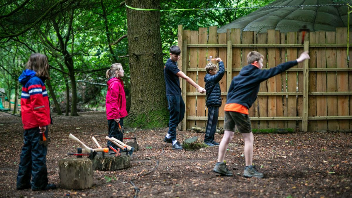 Children practicing axe throwing in a forest with a wooden fence backdrop.