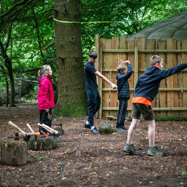 Children practicing axe throwing in a forest with a wooden fence backdrop.