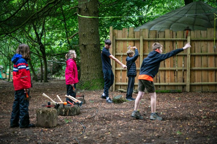 Children practicing axe throwing in a forest with a wooden fence backdrop.