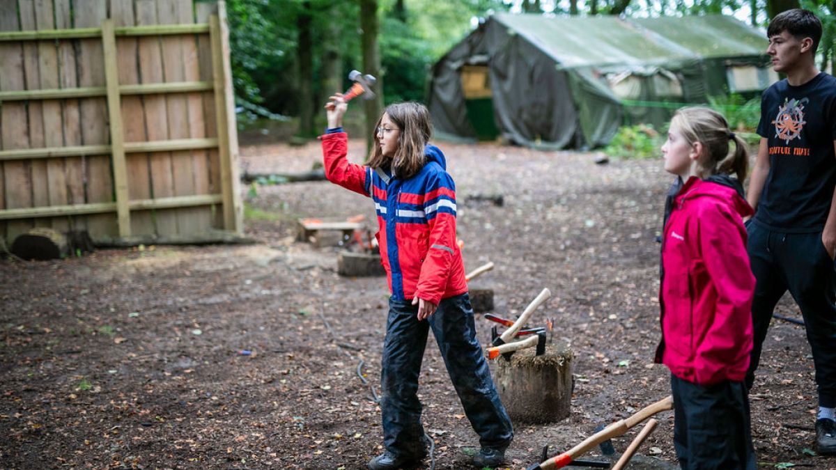 Child in red jacket throwing an axe at a target in wooded camp area.