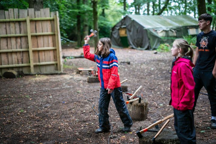 Child in red jacket throwing an axe at a target in wooded camp area.
