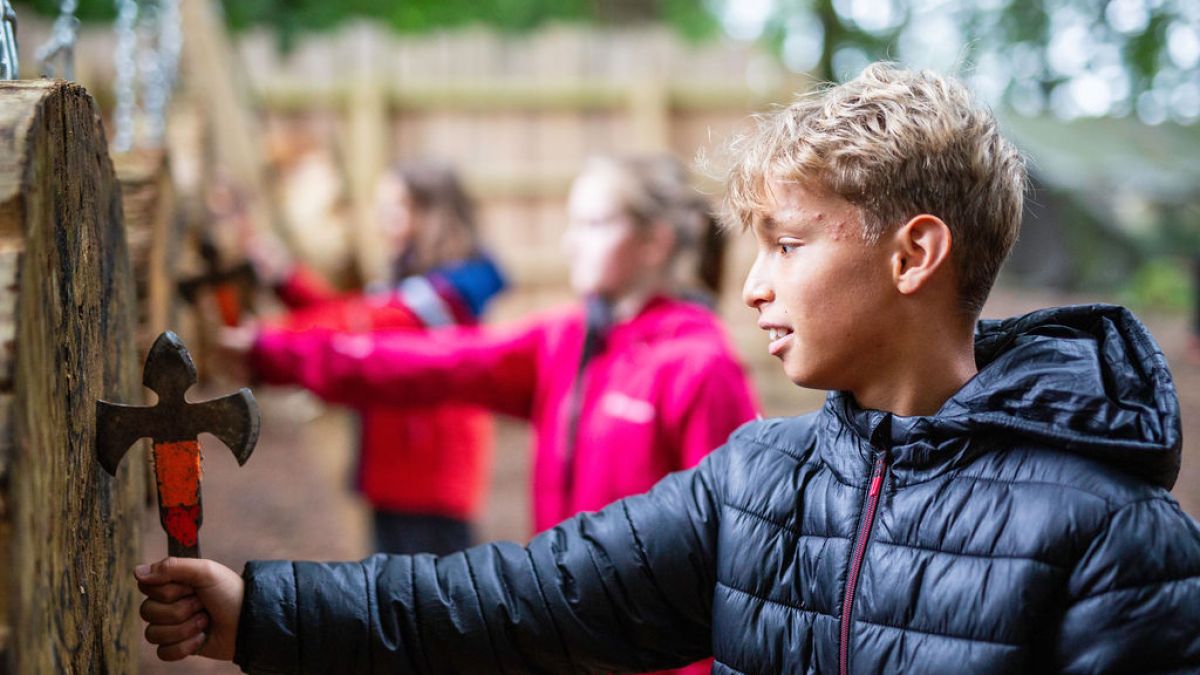 Boy in black jacket holds a throwing axe near wooden target outdoors.