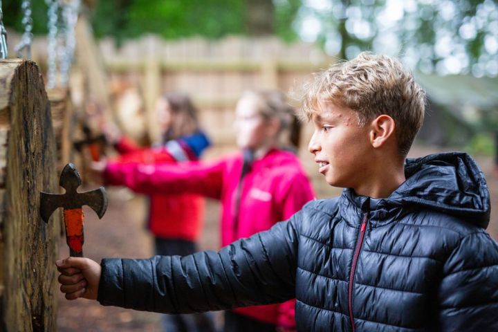 Boy in black jacket holds a throwing axe near wooden target outdoors.