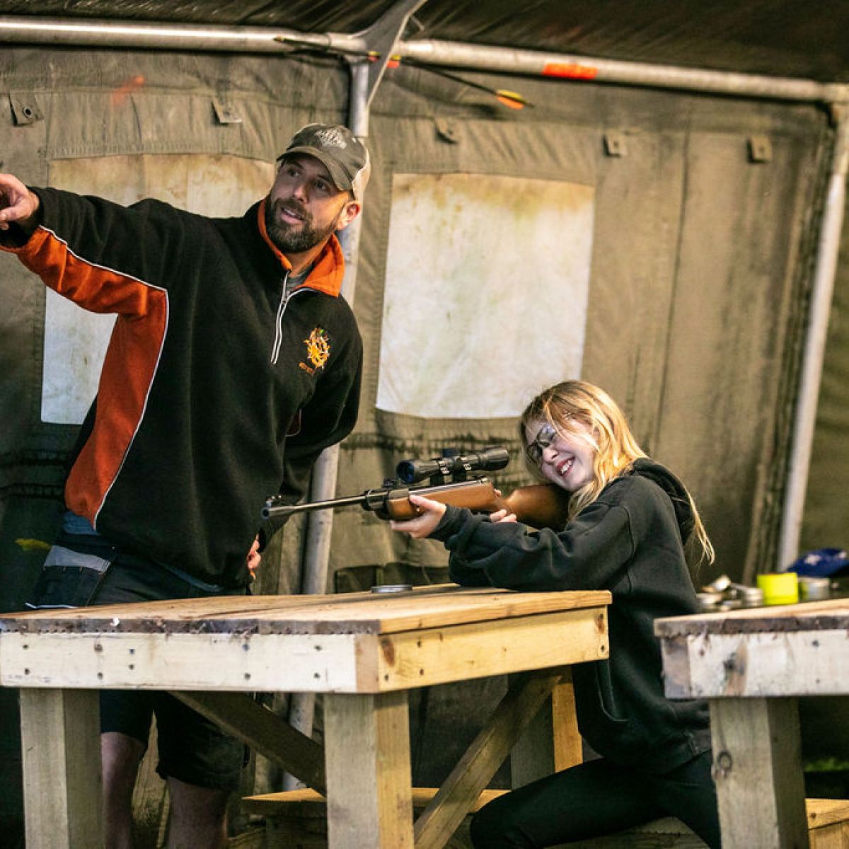 Person teaching girl to use a rifle at a wooden table in a tent.