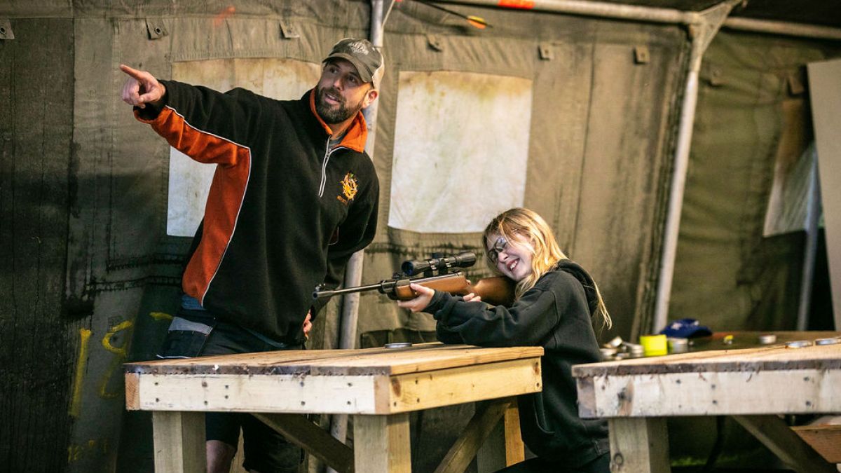 Person teaching girl to use a rifle at a wooden table in a tent.