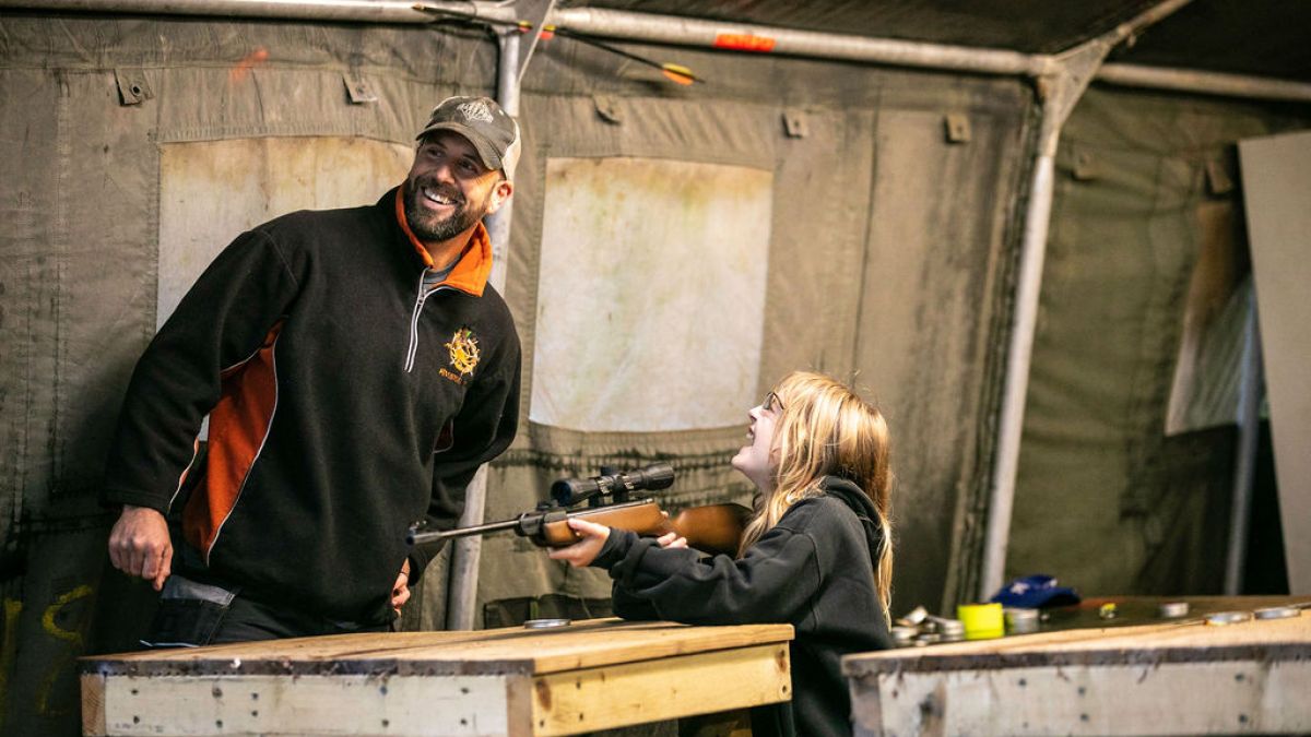 Man smiling while child aims rifle at a shooting range.