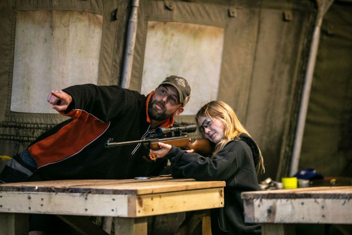 Man instructing child aiming rifle at wooden table in tent.