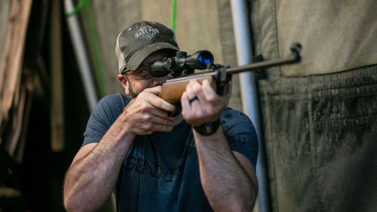 Man aiming a rifle with a scope indoors, wearing a cap and blue shirt.