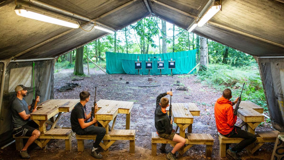 Four people sitting at wooden tables in a tent aiming rifles at targets in the woods.