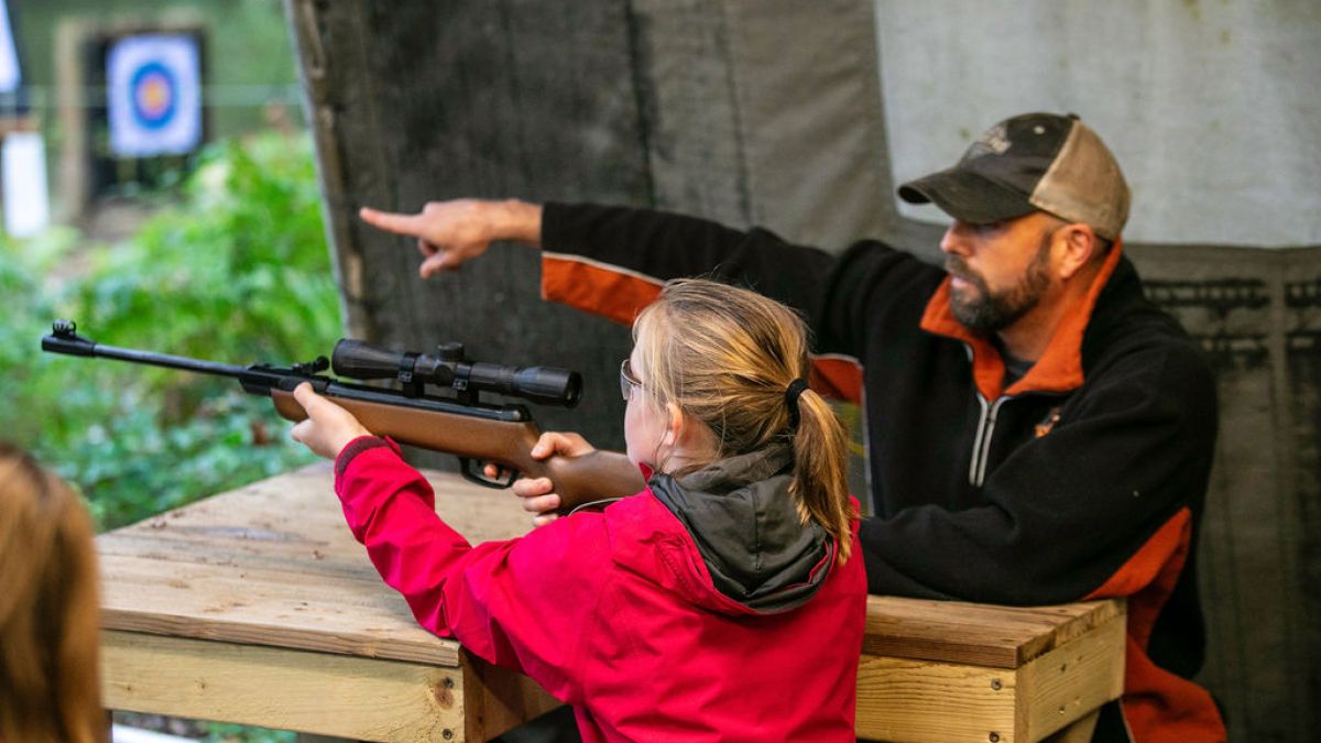 Child aims a rifle under adult supervision at a shooting range with a target in the background.