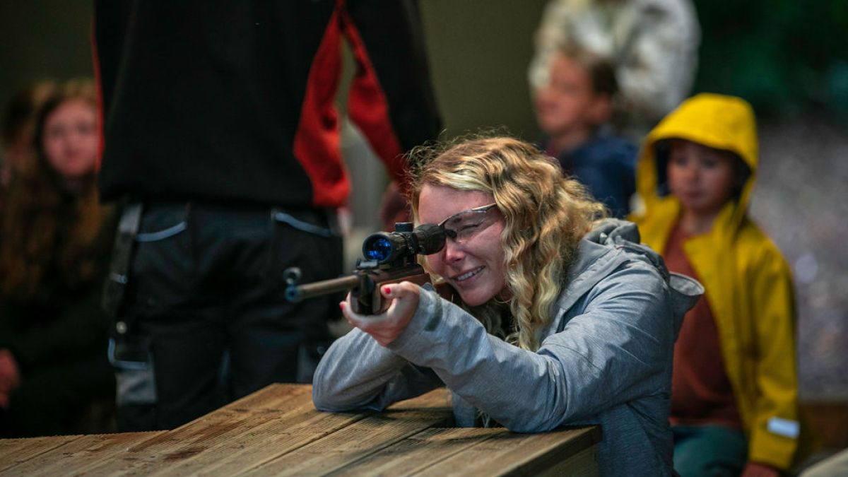 Person aiming rifle at shooting range table with onlookers in the background.
