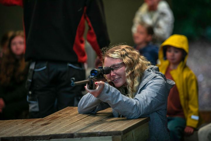 Person aiming rifle at shooting range table with onlookers in the background.