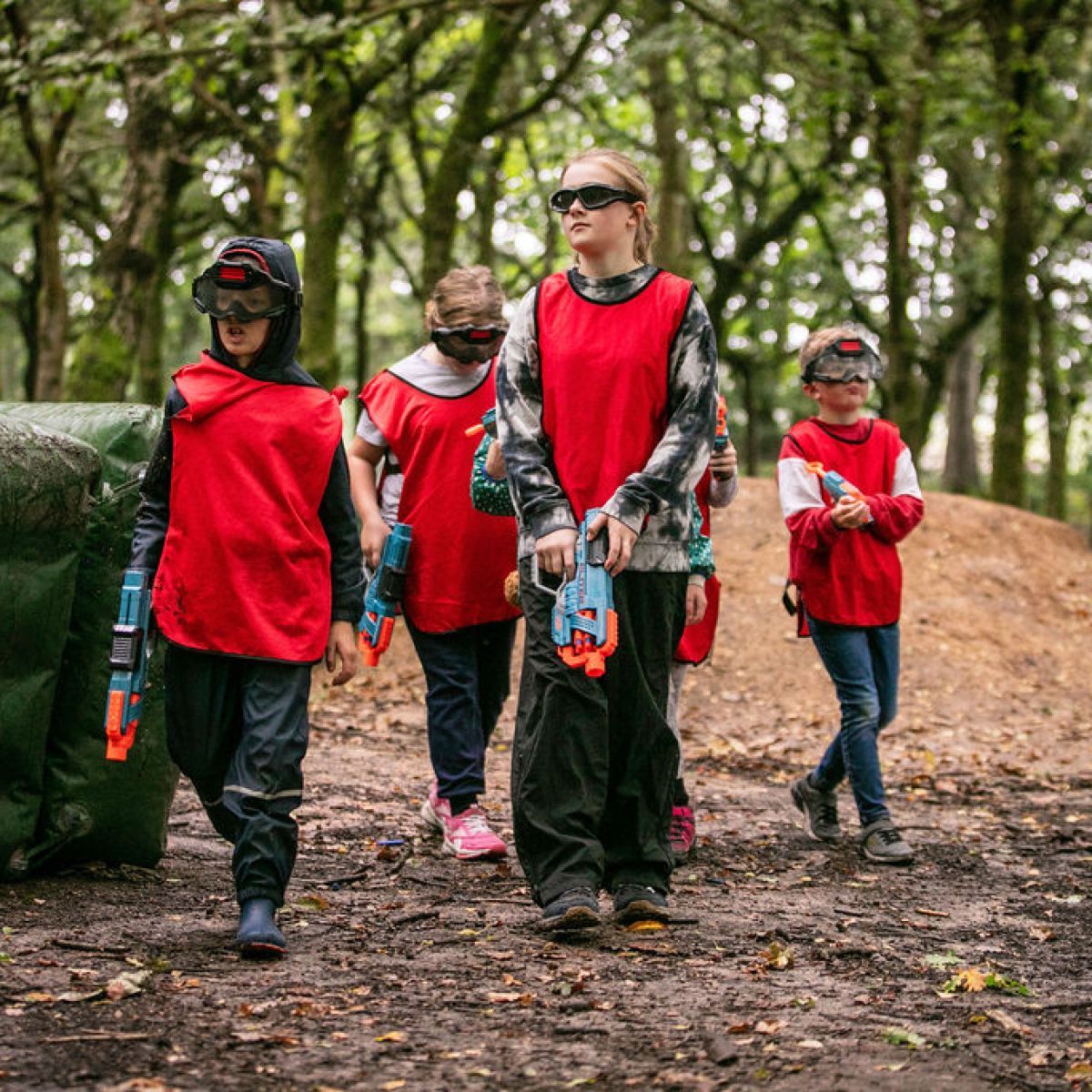 Children in red vests holding toy guns in a forest setting.