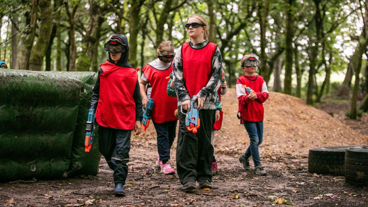Children in red vests holding toy guns in a forest setting.