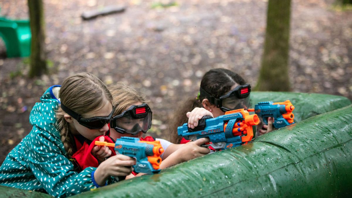 Three children outdoors playing with toy guns, wearing safety goggles.