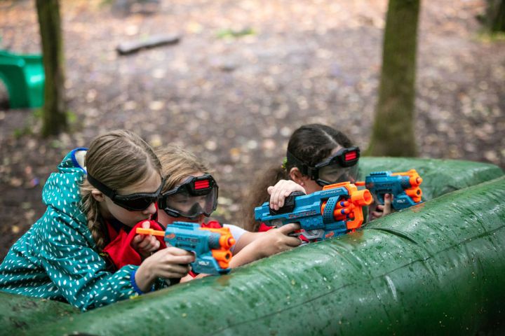 Three children outdoors playing with toy guns, wearing safety goggles.