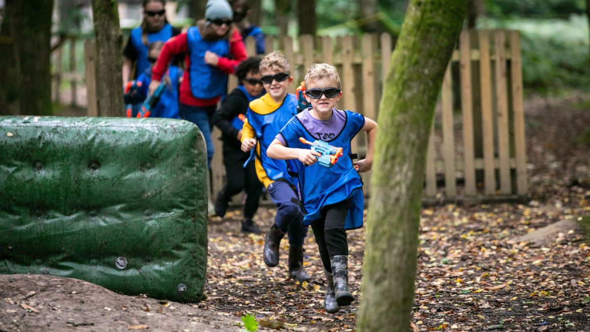 Children playing a Nerf game in the woods, wearing blue and yellow vests, running and holding toy guns.