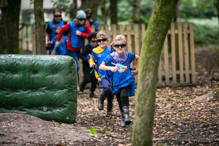 Children playing a Nerf game in the woods, wearing blue and yellow vests, running and holding toy guns.