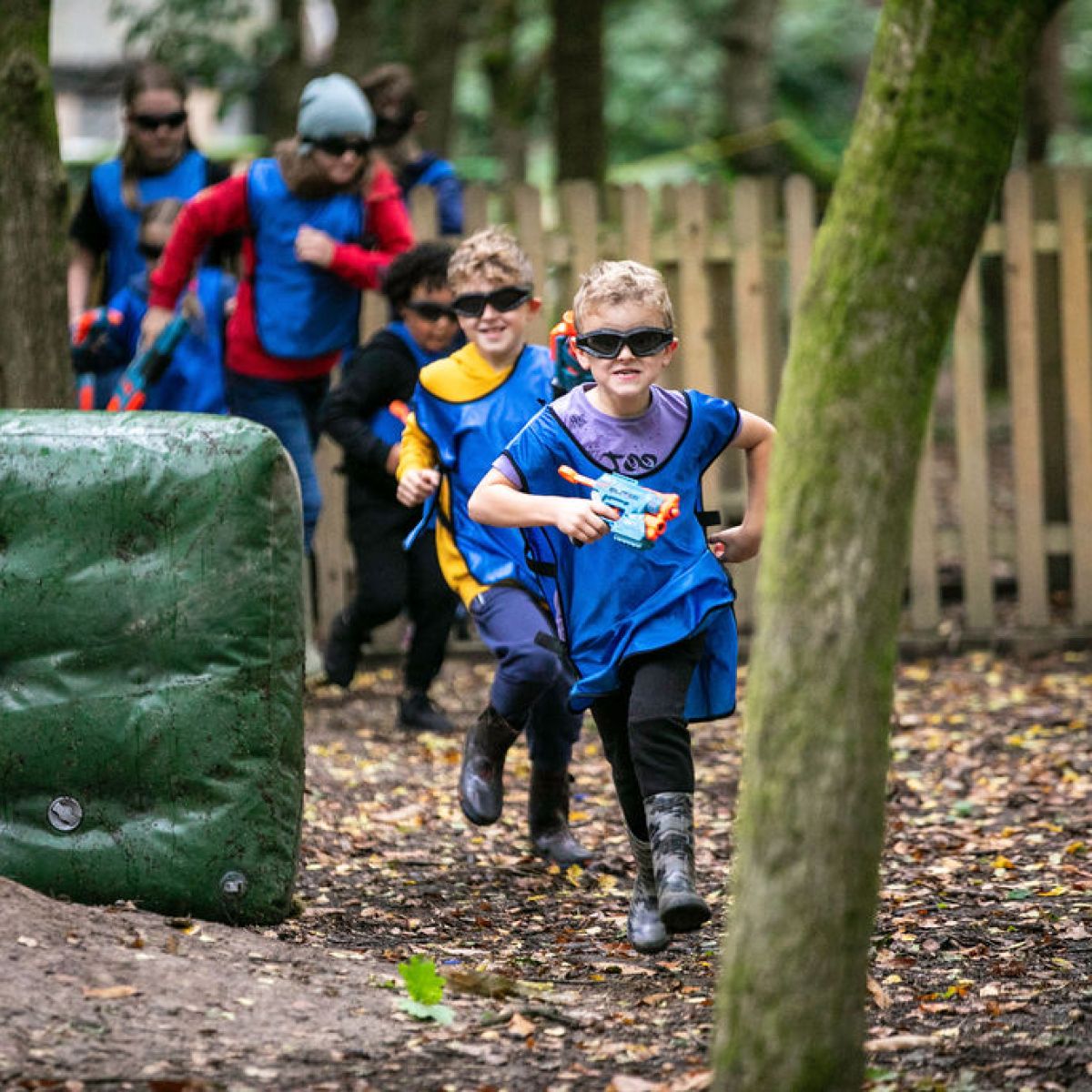 Children wearing blue and yellow vests run through a wooded area holding toy guns.