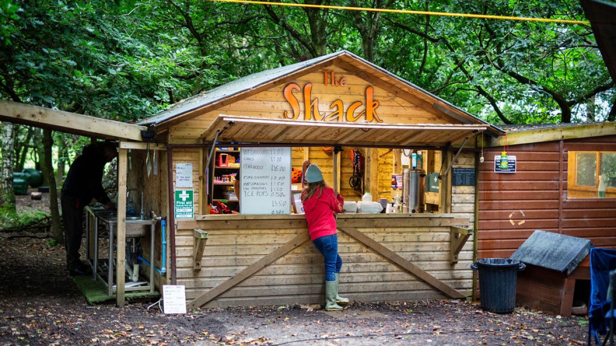 Person in red jacket at outdoor wooden shack with menu and trees.