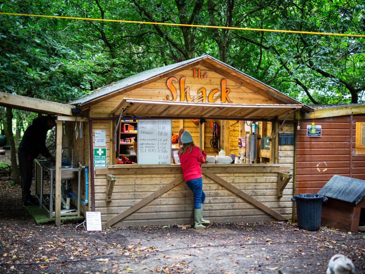 Person in red jacket at outdoor wooden shack with menu and trees.