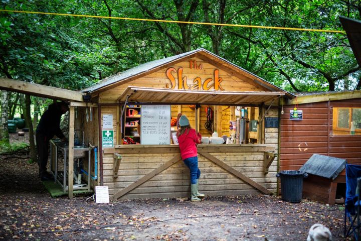 Person in red jacket at outdoor wooden shack with menu and trees.