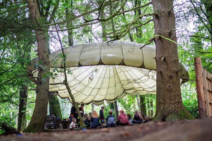 Children sit under a large canopy in a forest area with trees surrounding them.