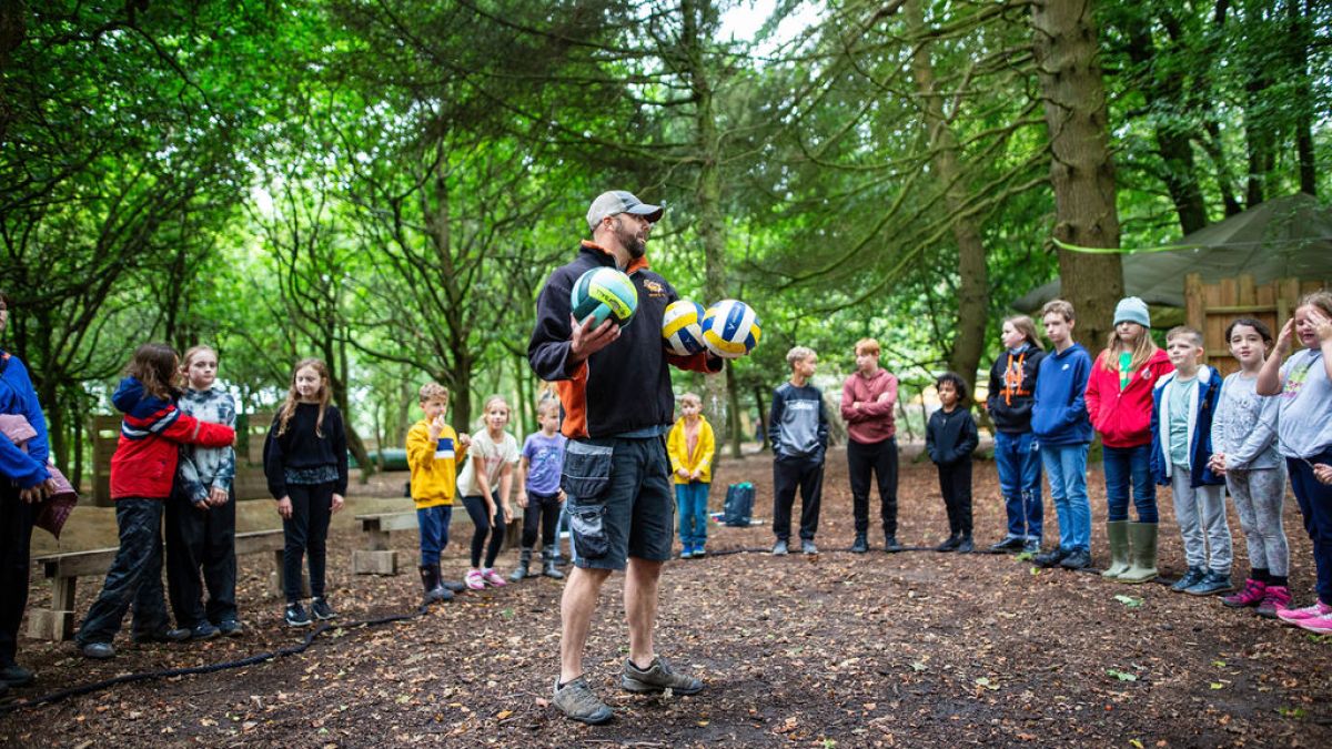Instructor holding volleyballs addresses kids in a forest outdoor setting.