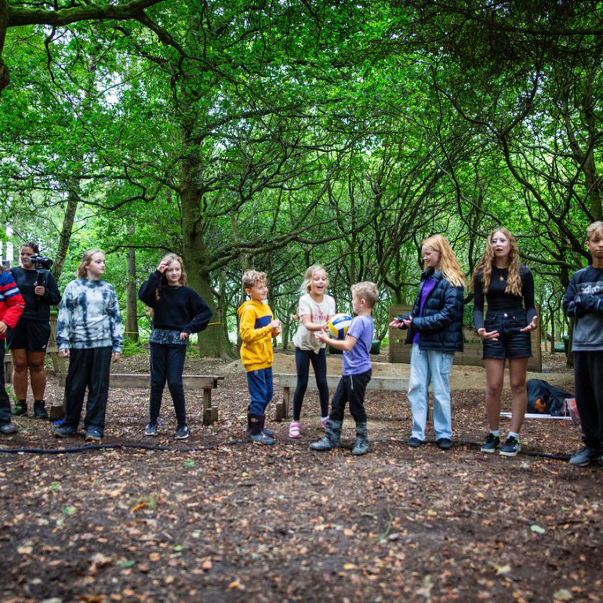 Group of kids standing in a forest clearing, some playing with a ball.