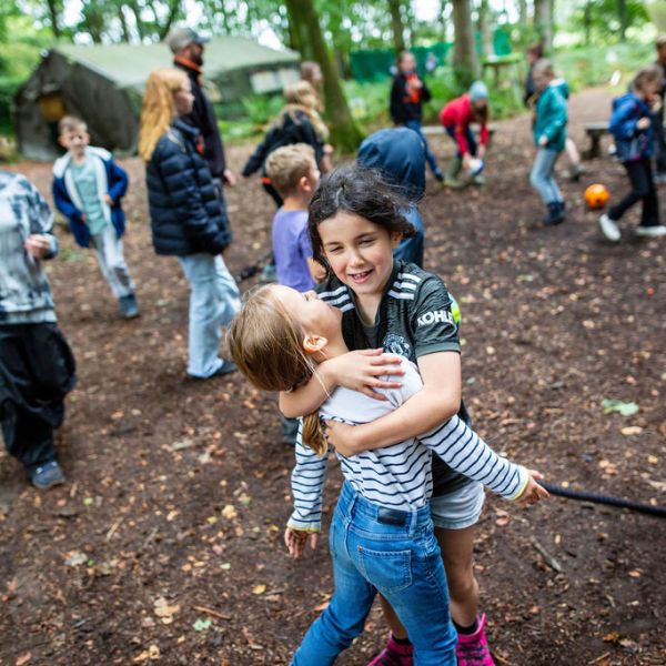Two children hugging outdoors with other kids playing in the background in a forest.