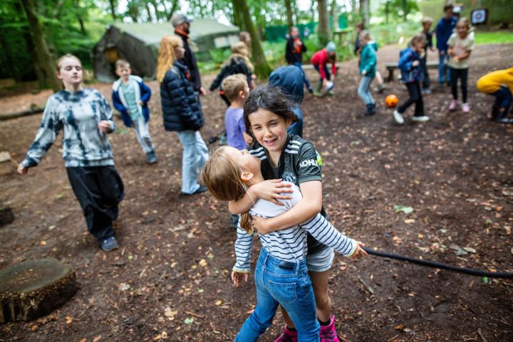 Two children hugging outdoors with other kids playing in the background in a forest.